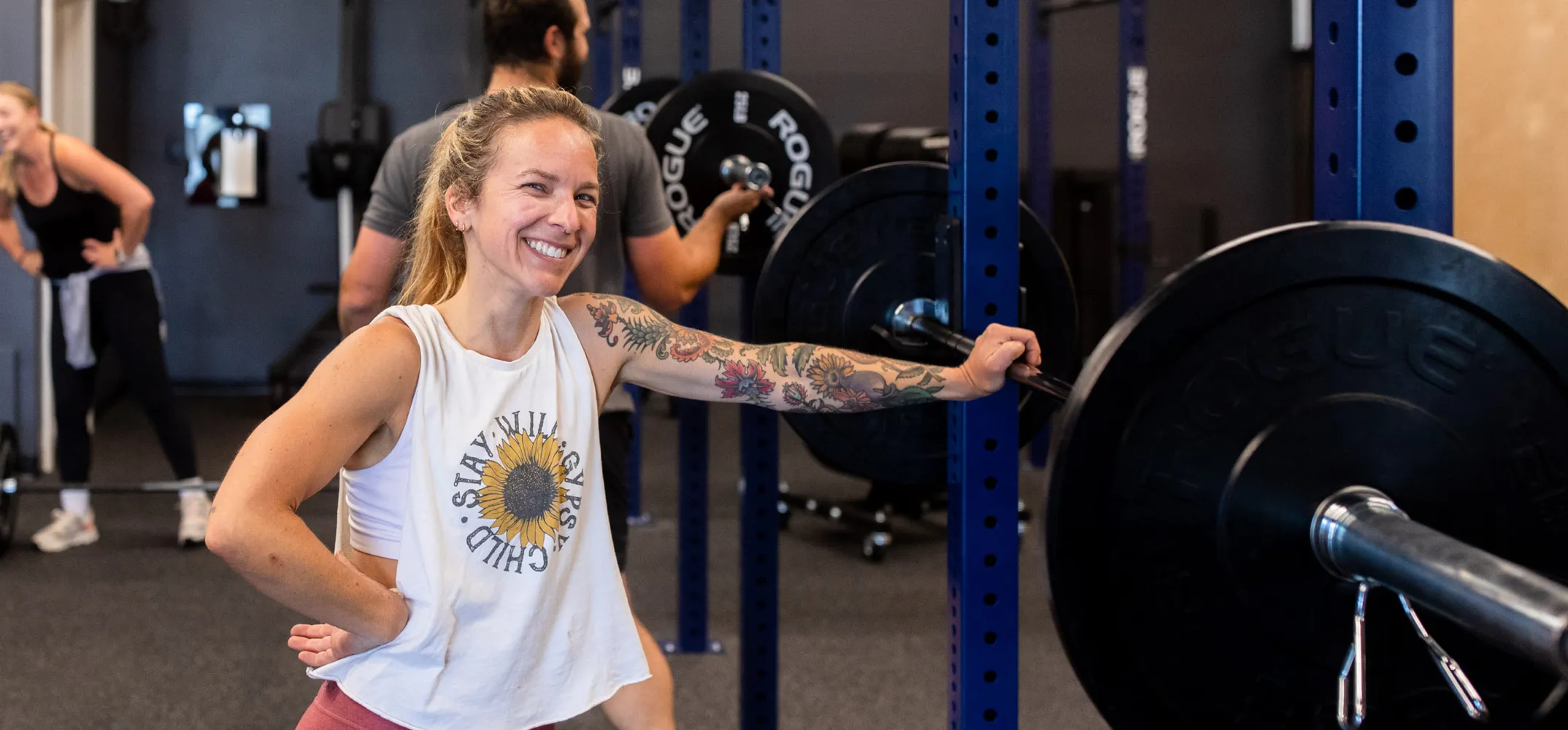 A smiling member with a floral tattoo sleeve resting on a barbell at a squat rack, with another member loading weight plates behind her at The Cove.