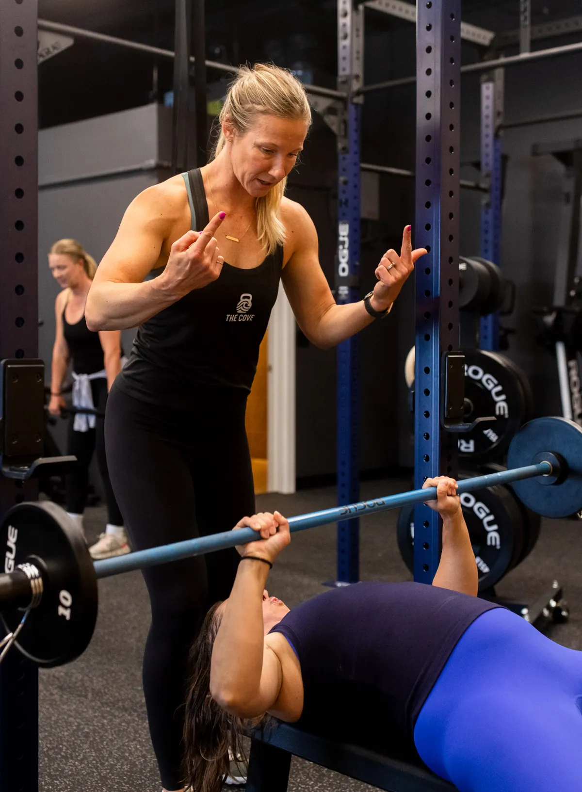 A Cove coach in a branded tank top coaching a member through a barbell bench press, with another member training in the background at The Cove.